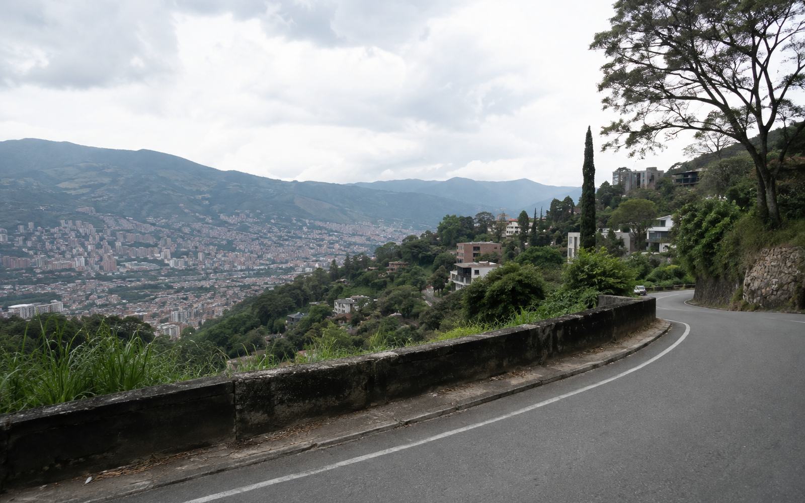 A curving hillside road high above the Aburrá Valley, houses stepping down the slope.